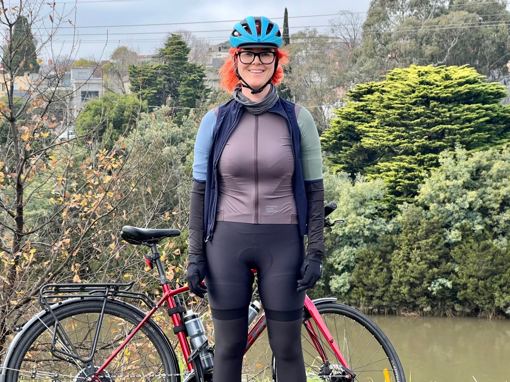 A person with red hair stands in front of a bike wearing a women's cycling outfit
